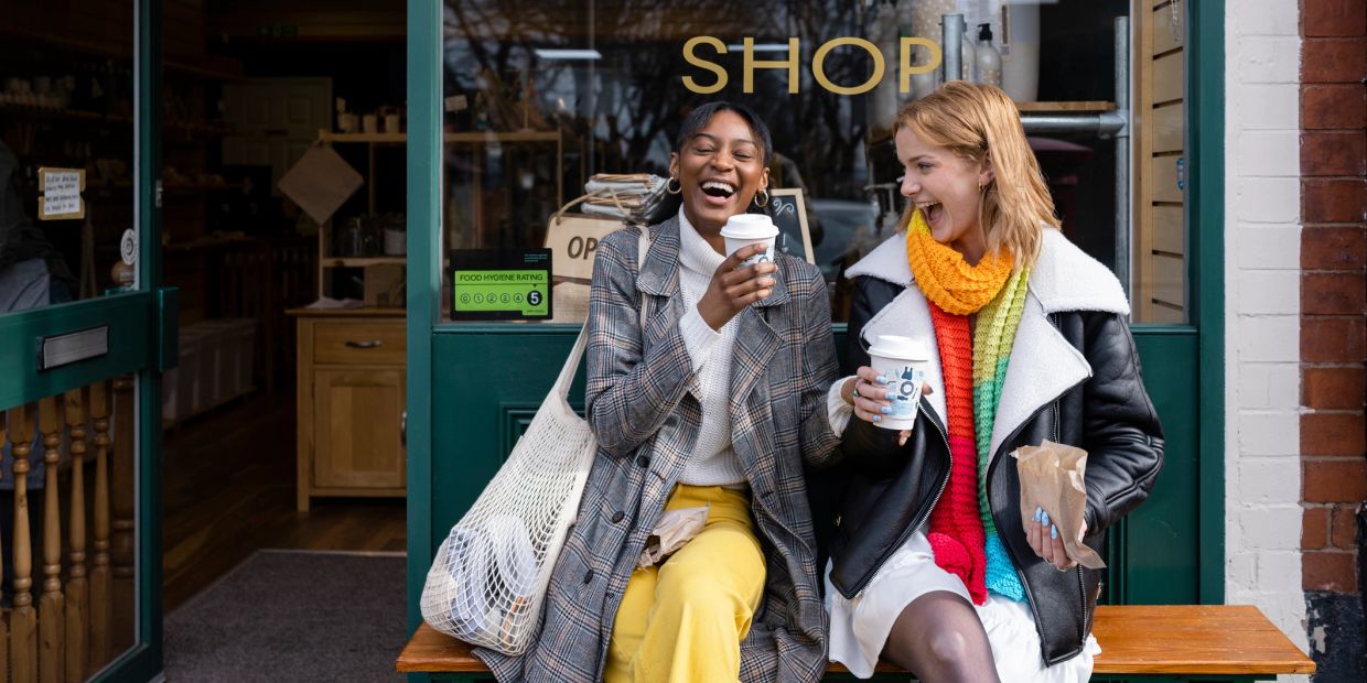 Two friends laughing and drinking coffee outside a shop.