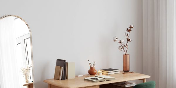 Minimalist wooden desk with books, vase, and cotton stems beside a tall mirror and curtain.