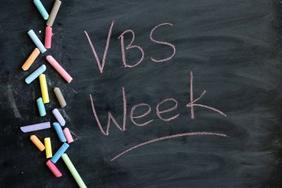 Colorful chalk sticks and VBS Week written on a blackboard.
