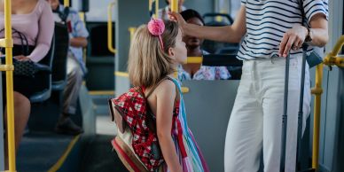 Little girl with backpack standing next to adult with suitcase on bus.