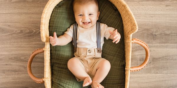 Smiling baby in beige suspenders lying in a woven basket on a wooden floor.