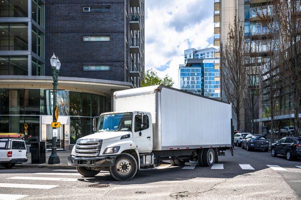 White delivery truck turning at an urban intersection with tall buildings.