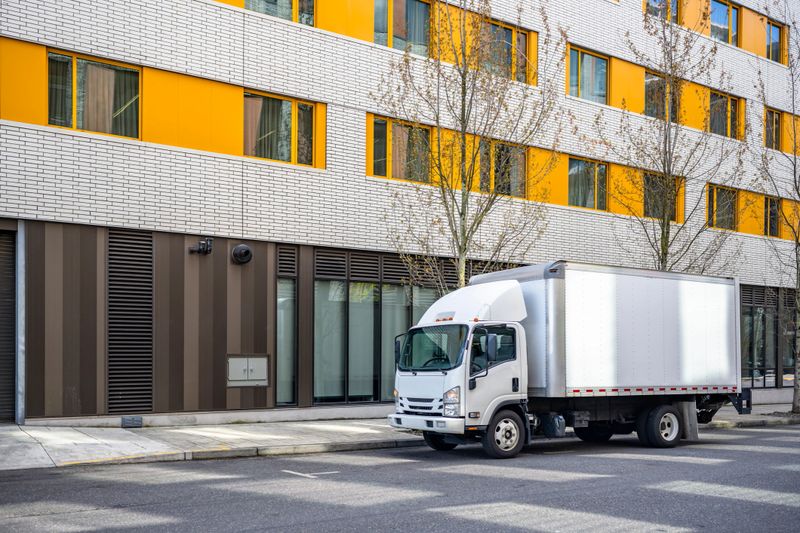 Small rig over cab white semi truck with long box trailer making local commercial delivery at urban city with multilevel residential apartments buildings standing on the street for upload cargo