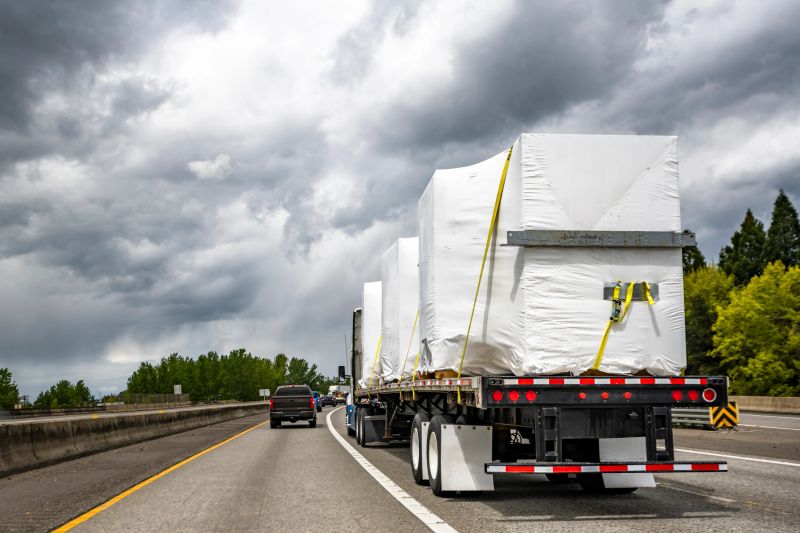 Blue and white big rig bonnet industrial semi truck tractor transporting covered fastened commercial cargo on flat bed semi trailer driving for delivery on the wide multiline turning highway road
