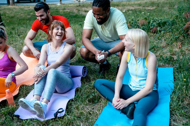 Multiethnic People after workout sitting on yoga mat on green grass in local Park. Happy smiling friends after yoga practice outdoors