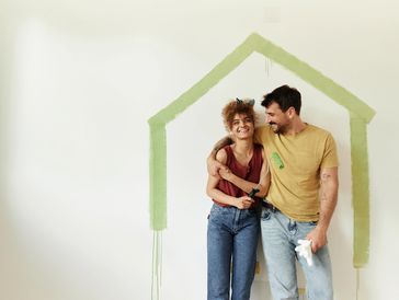 Couple painting a green house outline on a white wall, smiling and embracing.