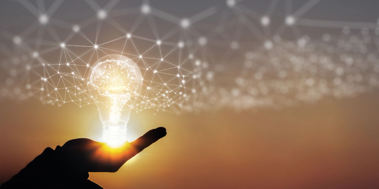 Hand holding a glowing lightbulb with a brain and network connections.