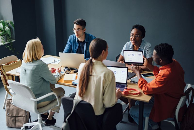 Serious multi ethnic group of friends talking and analyzing graph for school project using a laptop computers and digital tablet while sitting at desk together.