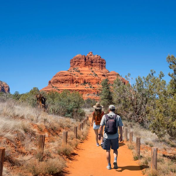 Two hikers walking on a trail towards red rock formations.