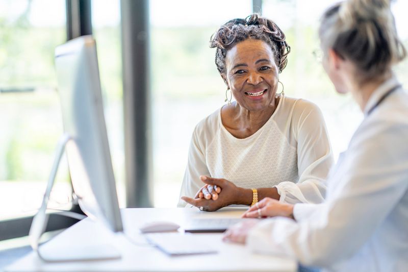 A senior woman of African decent, sits with her young doctor at her desk as they meet to discuss her health.  The woman is dressed casually and smiling at the doctor as she listens attentively.  The doctor is holding a tablet as she takes notes electronically for the woman's file.