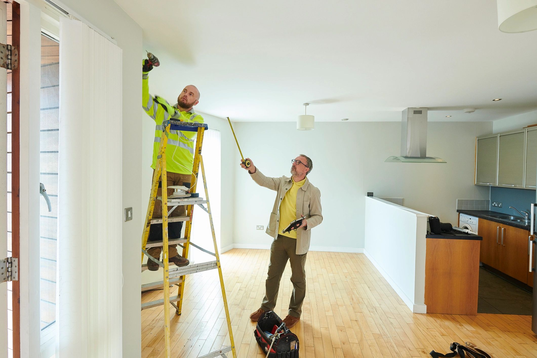 A worker on a ladder installs a fixture while another measures in a modern kitchen.