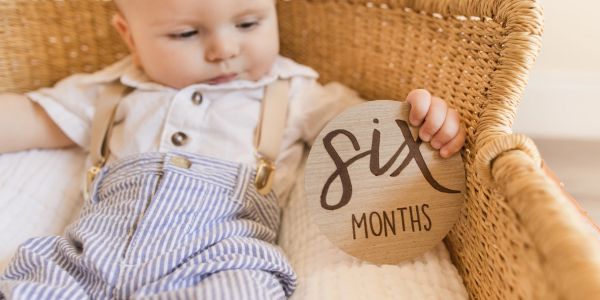 Baby holding a "six months" milestone plaque in a basket.