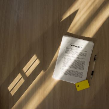Contract, pen, and yellow card on wooden floor with light shadows.