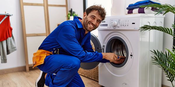 A repairman in a blue jumpsuit kneels repairing a washing machine