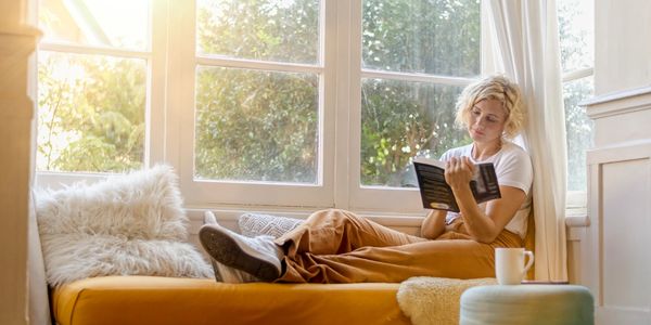 A woman reads a book by a sunlit window on a cozy daybed.