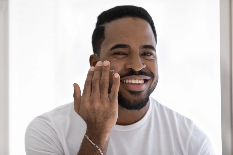 Handsome smiling African man applying moisturizer facial creme, looks at camera feels satisfied. Nourishing anti-wrinkles treatment for soft smooth hydrated skin, boost elasticity. Skincare concept