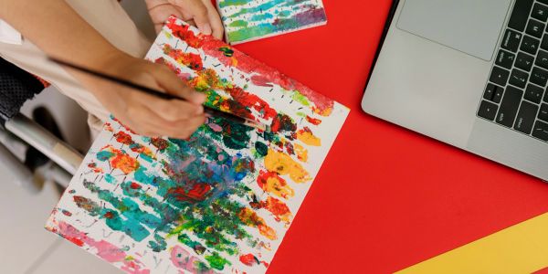 Person painting colorful abstract art on paper next to a laptop.
