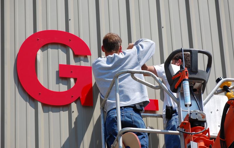 A pair of workers install a sign on the side of an industrial building.