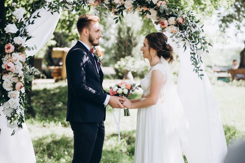 Stylish groom in a suit and beauty bride in a white dress in front wedding arch decorated with flowers. Wedding portrait of the newlyweds.