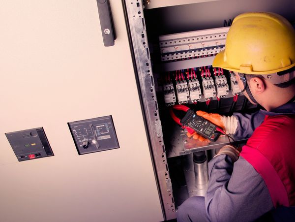 Electrician testing electrical panel with a multimeter wearing safety gear.