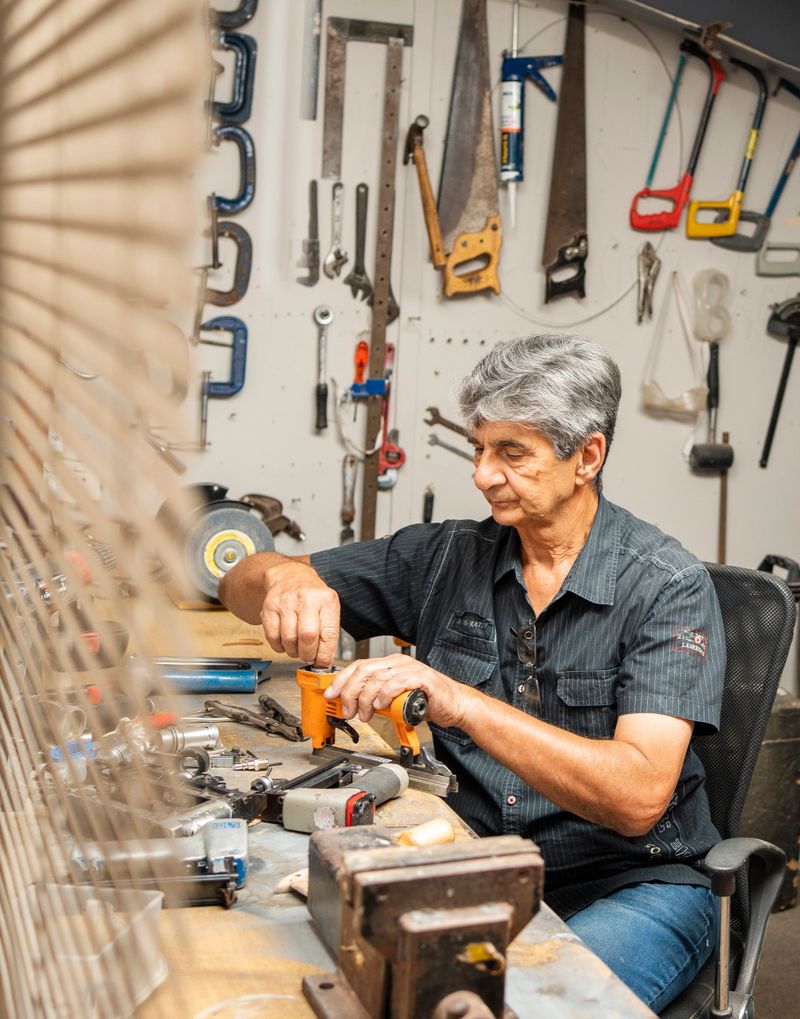Mature man repairing something while sitting at a table surrounded by tools in a woodworking workshop
