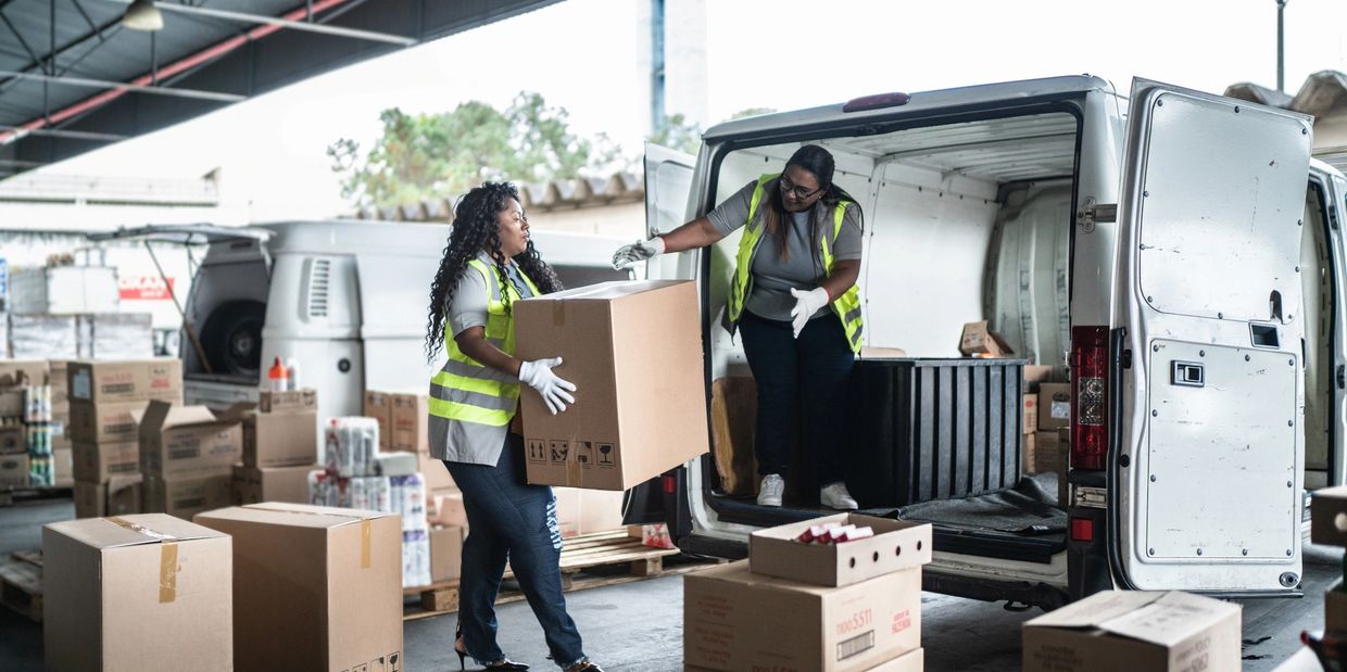 Two women unloading boxes from a van at a warehouse.
