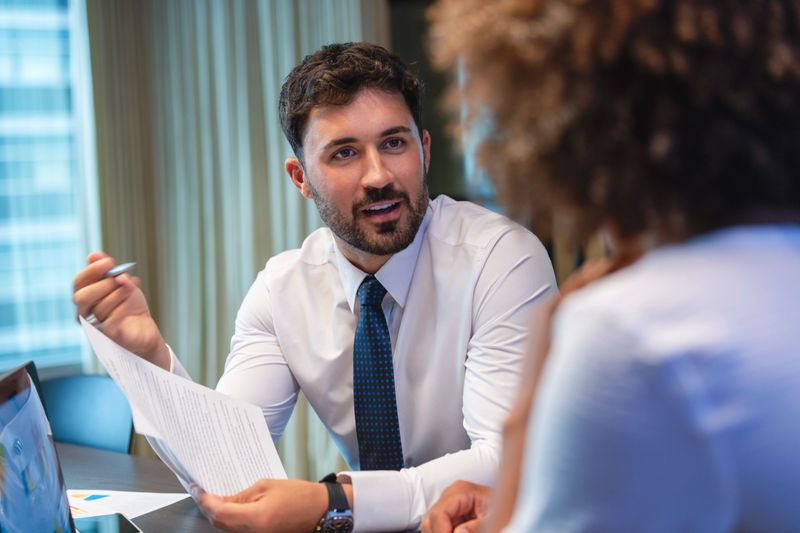 Business man talking to a business woman while holding a document. He is happy and smiling. He is wearing a tie.