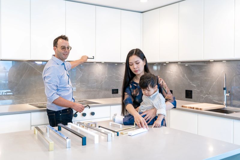 Caucasian male interior designer  demonstrating cabinet and door handle pulls for kitchen renovation remodel. East Asian female mother makes selection while holding young, curious boy child.