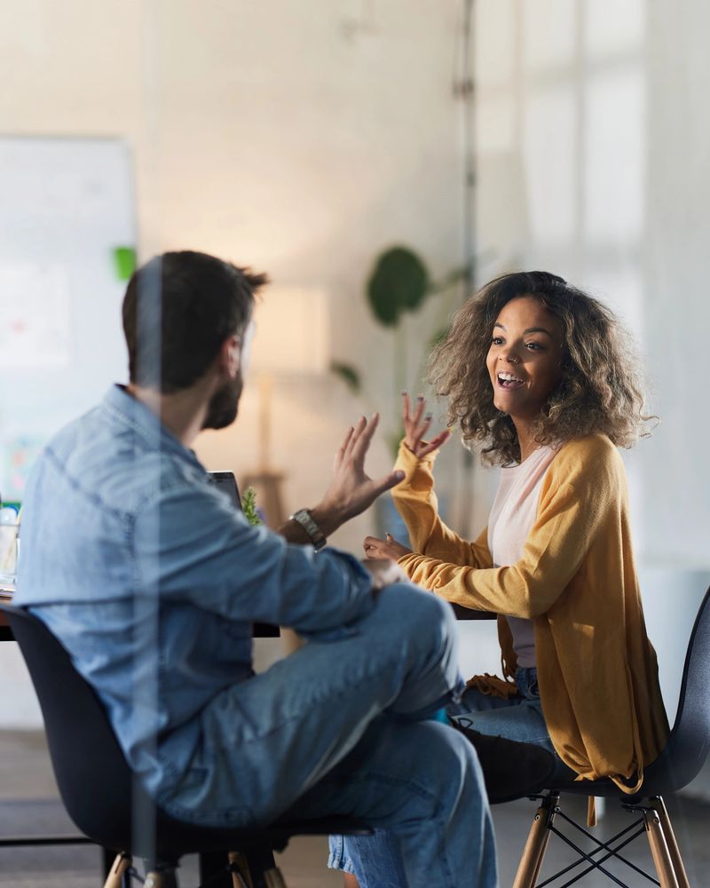 Happy creative colleagues talking while having a meeting at casual office. Focus is on woman. The view is through glass.