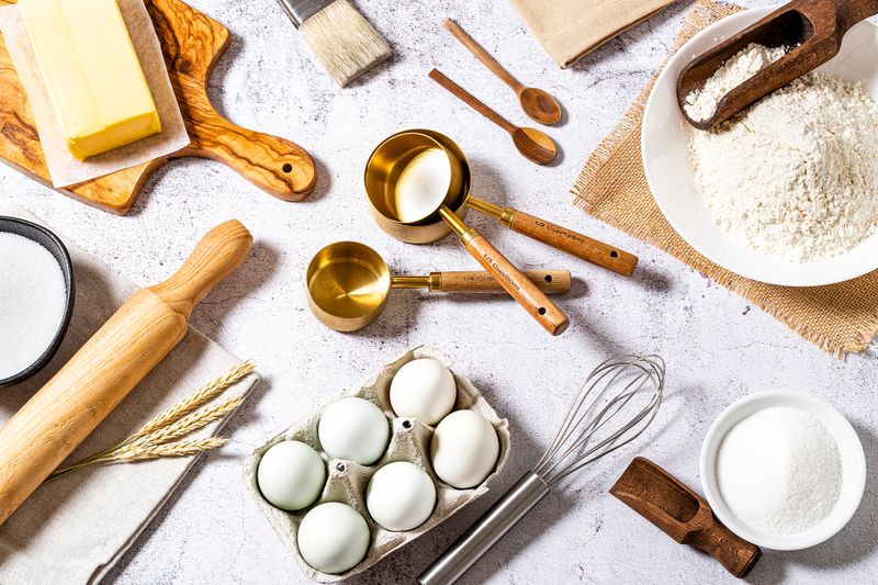 Top view of various bakery  ingredients and items such as eggs, butter, flour, sugar, rolling pin, measure cups, hand whisk and serving scoop disposed all around the image on a bakery concept background