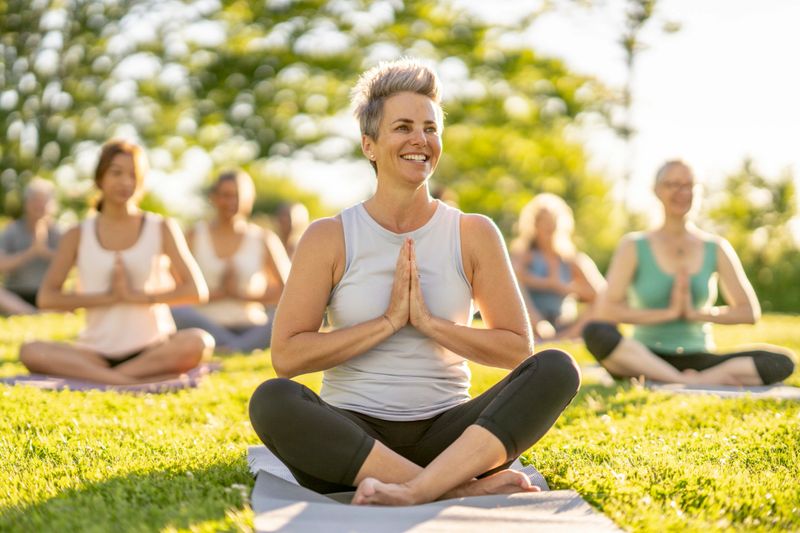 A large group of adults sit on yoga mats in the grass on a sunny summers evening.  They are each wearing comfortably athletic wear and are cross-legged with their hands on their knees in a prayer pose as they participate in a class together.
