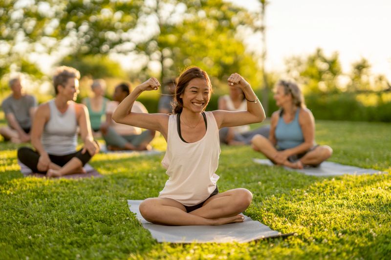 A woman sits on a yoga mat in the grass on a sunny summers evening as she flexes her muscles.  She is seated among a group of adults as they participate in a fitness class together.  They are each dressed comfortably in athletic ear and smiling.