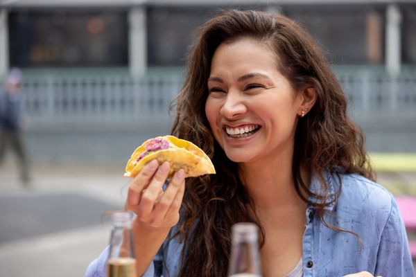 A joyful woman holding two tacos, smiling brightly outdoors.