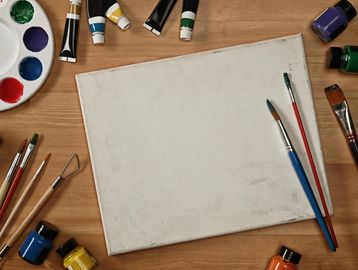 Blank canvas surrounded by various paintbrushes, tubes, and jars of paint on a wooden table.