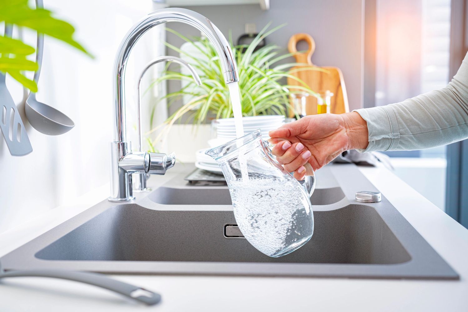 Hand filling a glass pitcher with water from a kitchen faucet.