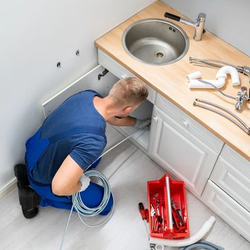 Plumber fixing pipes under kitchen sink with tools and parts nearby.