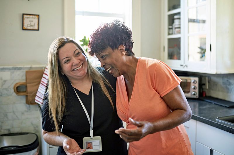 Waist-up view of Black client and healthcare worker standing with arms around each other and laughing during routine house call.