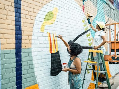 Two women painting a colorful mural on a brick wall.