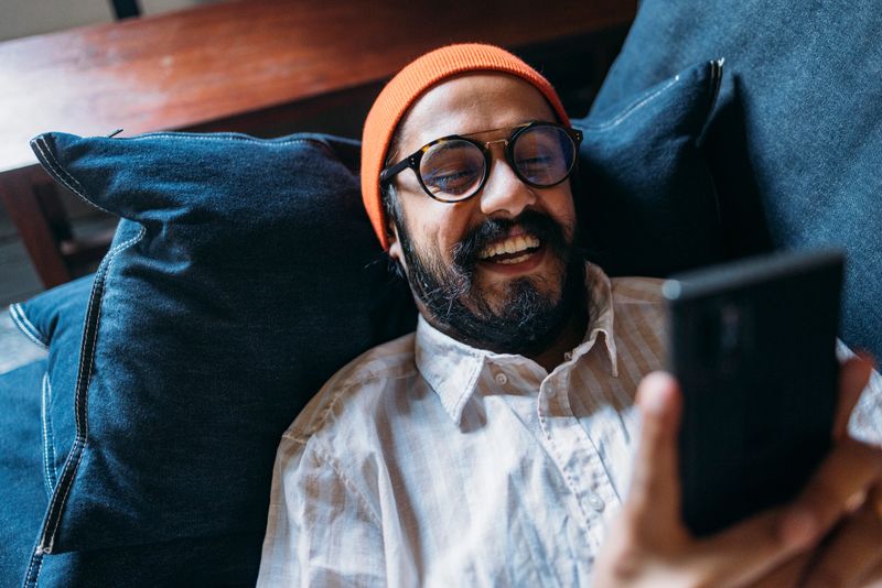 A from above view of a smiling Indian influencer fashionably dressed spending his leisure time relaxing using his smartphone.