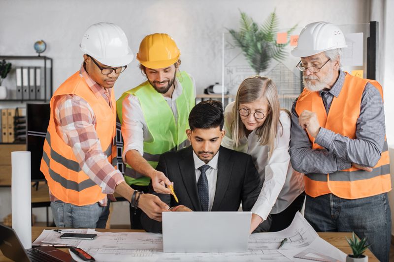 Teams of architects, builders and designers working on project of new modern house. The head of company sits at table and shows on laptop sketches of building while contractors standing around.