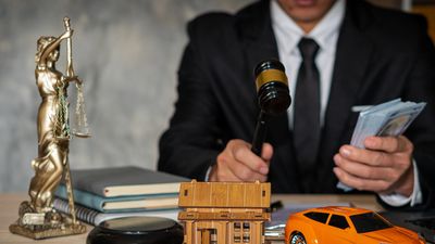 Judge holding money with legal symbols, house model, and toy car on the table.