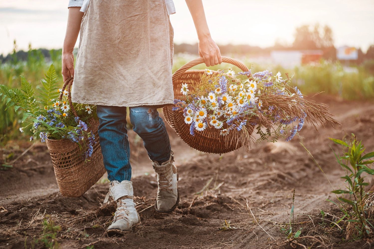 Person carrying baskets of wildflowers and ferns in a rustic field.