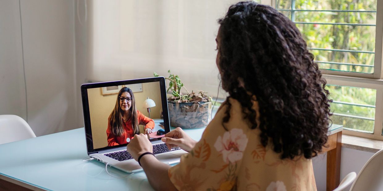 Woman video calls another woman on a laptop at a glass table in a bright room.