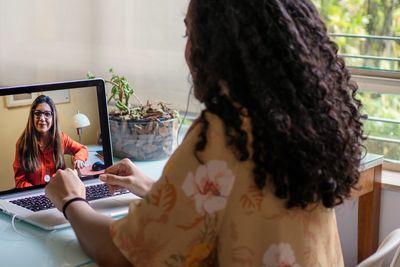 Woman video calls another woman on a laptop at a glass table in a bright room.