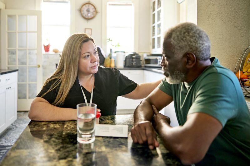 Waist-up view of Latin American home caregiver sitting at kitchen table with Black man in early 70s discussing plans for wellness management.