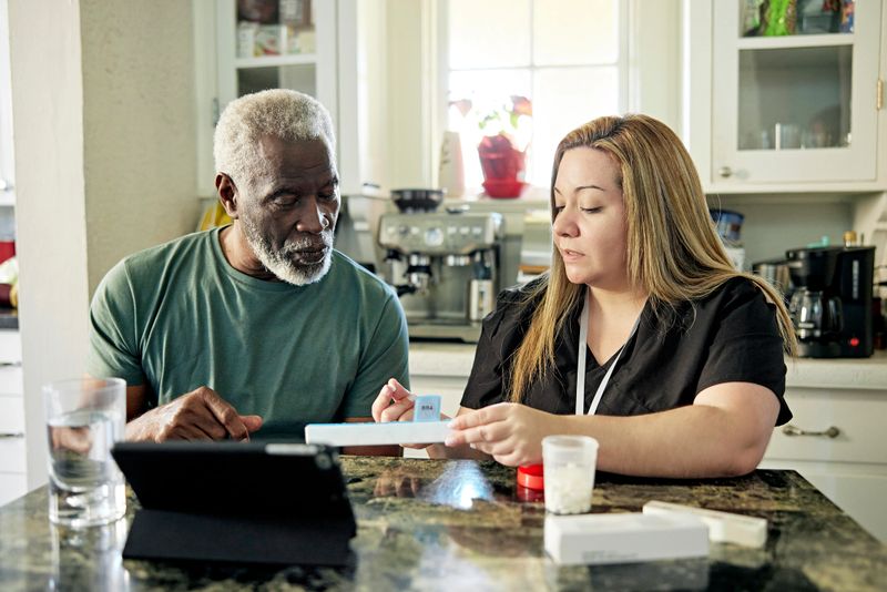 Front view of early 70s Black man sitting at kitchen table next to healthcare worker instructing him about what medication to take and when.