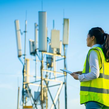 Engineer in a high-visibility vest inspects telecom antennas using a tablet outdoors.