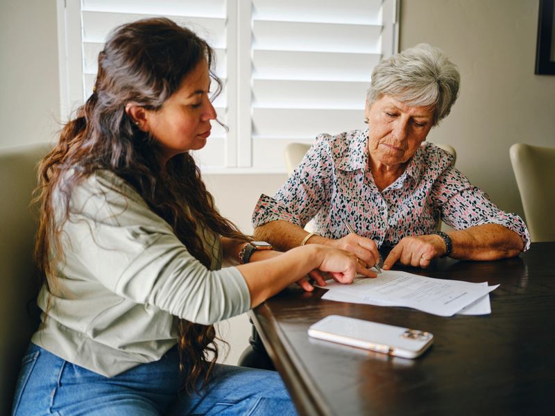 A Hispanic woman, helping a senior aged woman prepare financial documents.