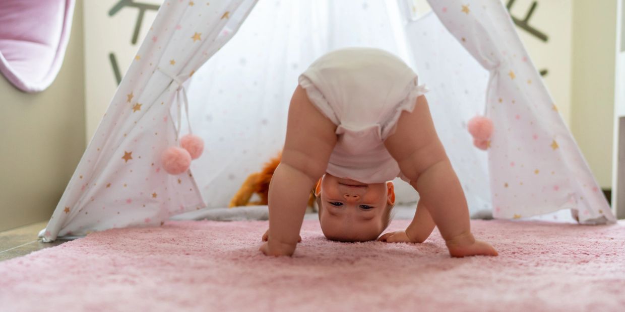 A baby playfully looks upside down inside a starry play tent on a pink rug.