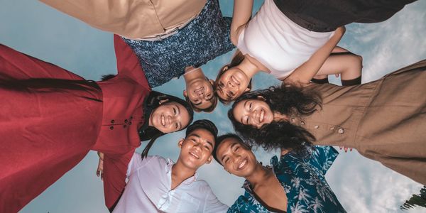 Group of six friends smiling in a circle, arms around each other, looking down at the camera.